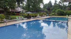 Una piscina con solárium de baldosas, rodeada de sillas y sombrillas verdes, y un edificio con techo inclinado al fondo.