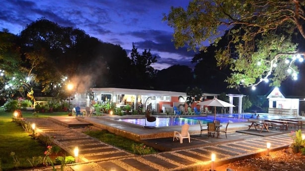 Un espace extérieur avec piscine, aménagé pour la nuit, avec un chemin en pierre, des chaises longues et un bâtiment en arrière-plan.