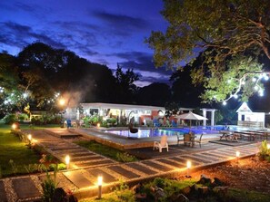 Un área de piscina exterior de noche con un sendero de piedra, sillas de descanso y un edificio al fondo.
