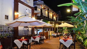 Een terras met tafels en stoelen, overdekt met parasols, en een gebouw met een traditionele architectuur op de achtergrond.