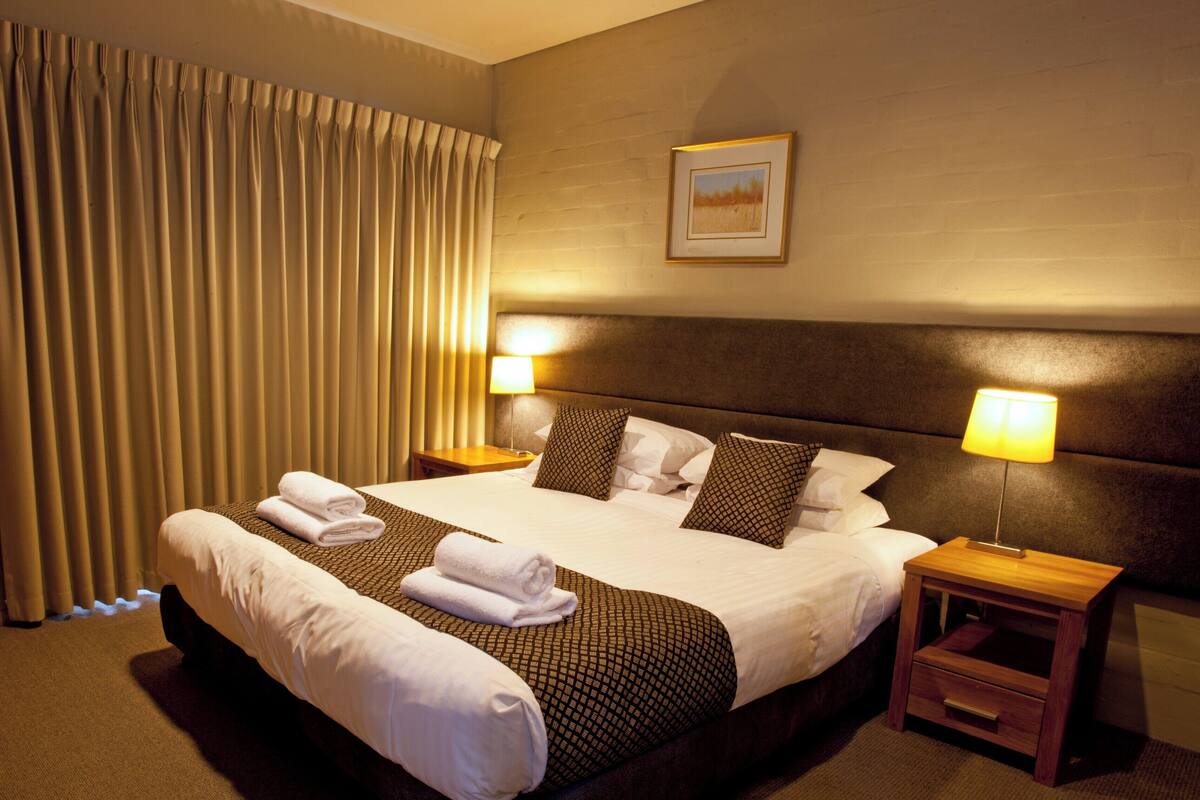 A hotel room with a large bed, two bedside lamps, a headboard, a framed picture on the wall, and a window with curtains.