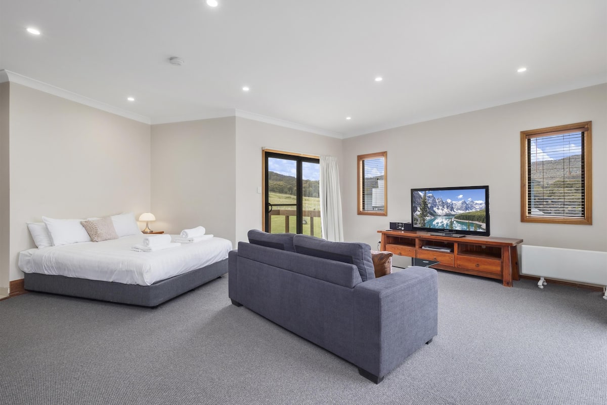 A modern hotel room with a bed, grey sofa, TV, and a scenic view through the windows.