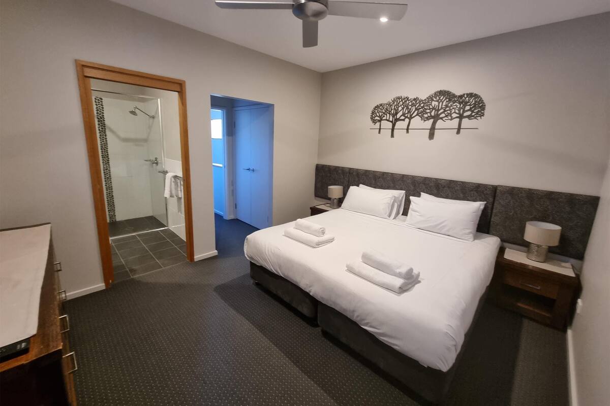 A hotel room with a bed, a ceiling fan, a nightstand, a wall art of trees, and a bathroom with a shower and a towel rack.