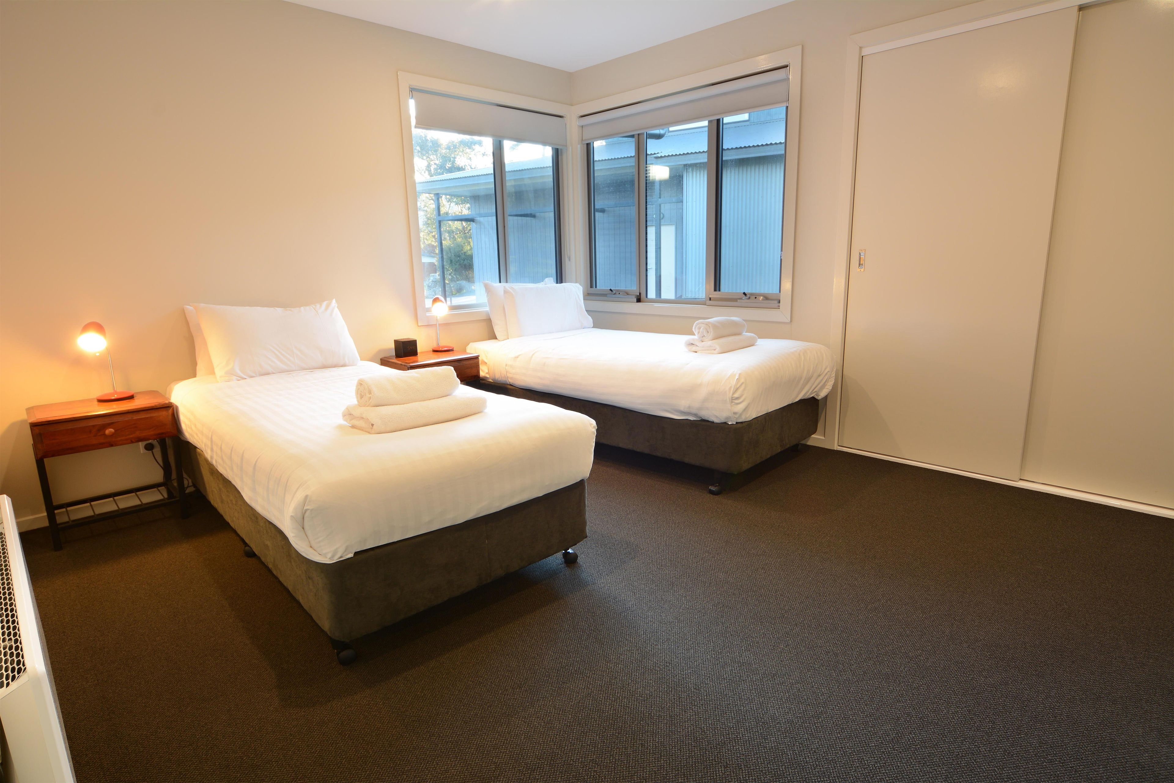A hotel room with two beds, a nightstand, a lamp, and a window with blinds.
