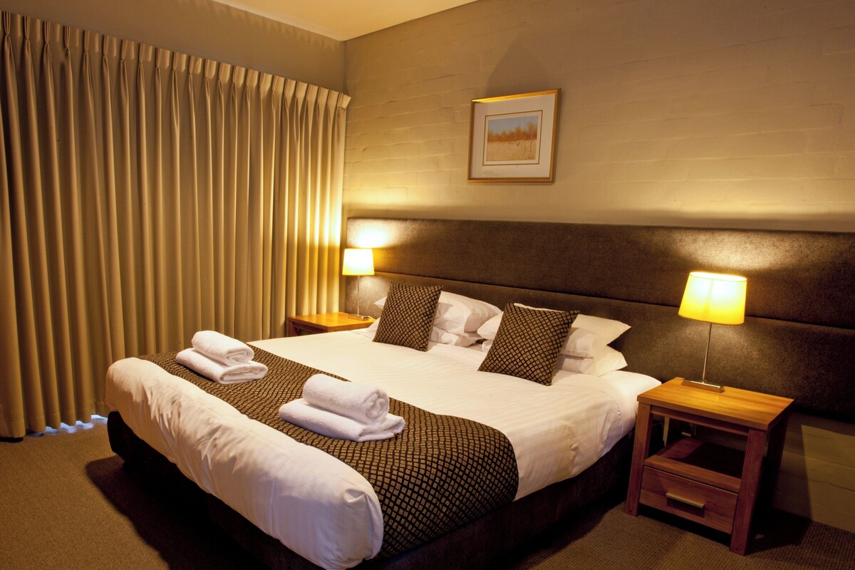 A hotel room with a large bed, two bedside tables, a headboard, a framed picture on the wall, and a window with curtains.