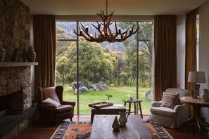 Une pièce dotée d’une cheminée en pierre, de fauteuils en cuir, d’une table basse en bois et d’une grande fenêtre donnant sur un paysage boisé.