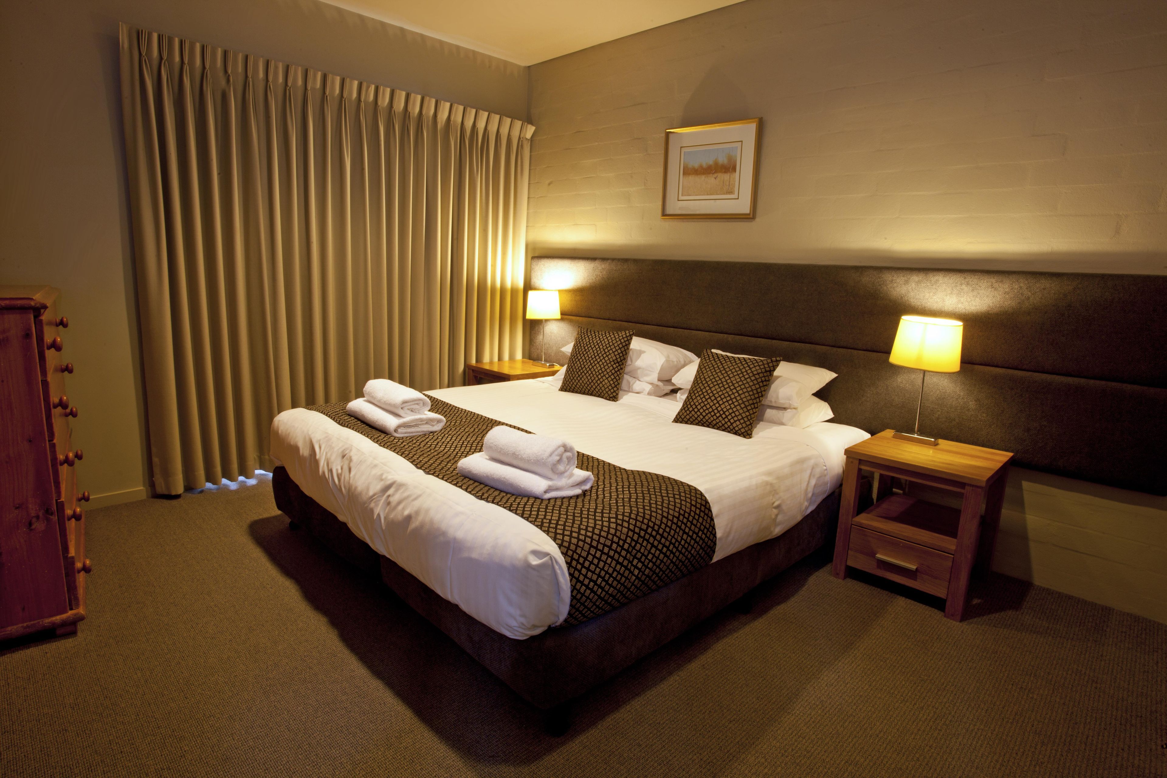 Quarto de hotel com uma cama grande, duas mesas de cabeceira, cabeceira, um quadro na parede e uma janela com cortinas.