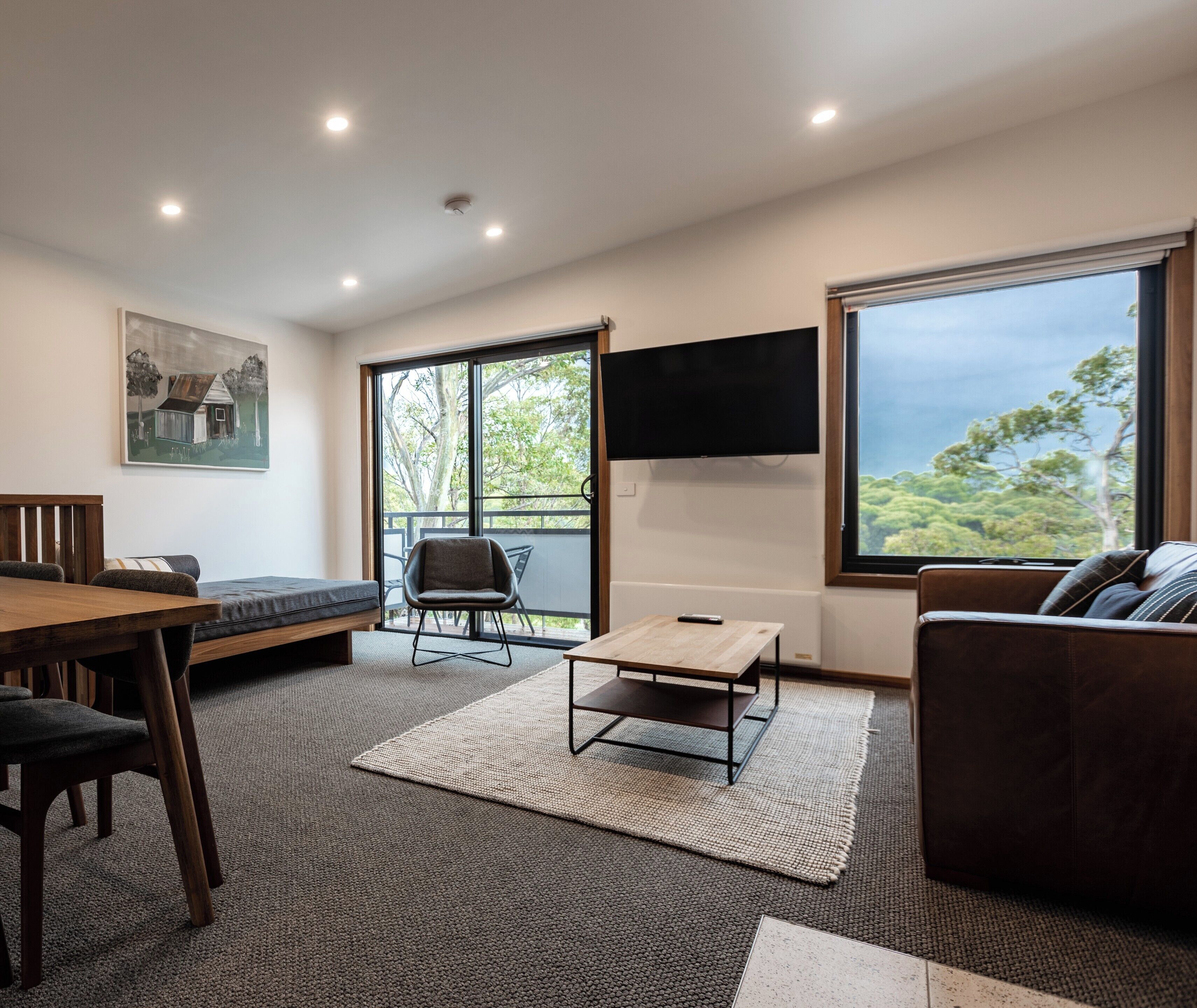 A modern living room with a wooden dining table, a sofa, and a TV mounted on the wall.