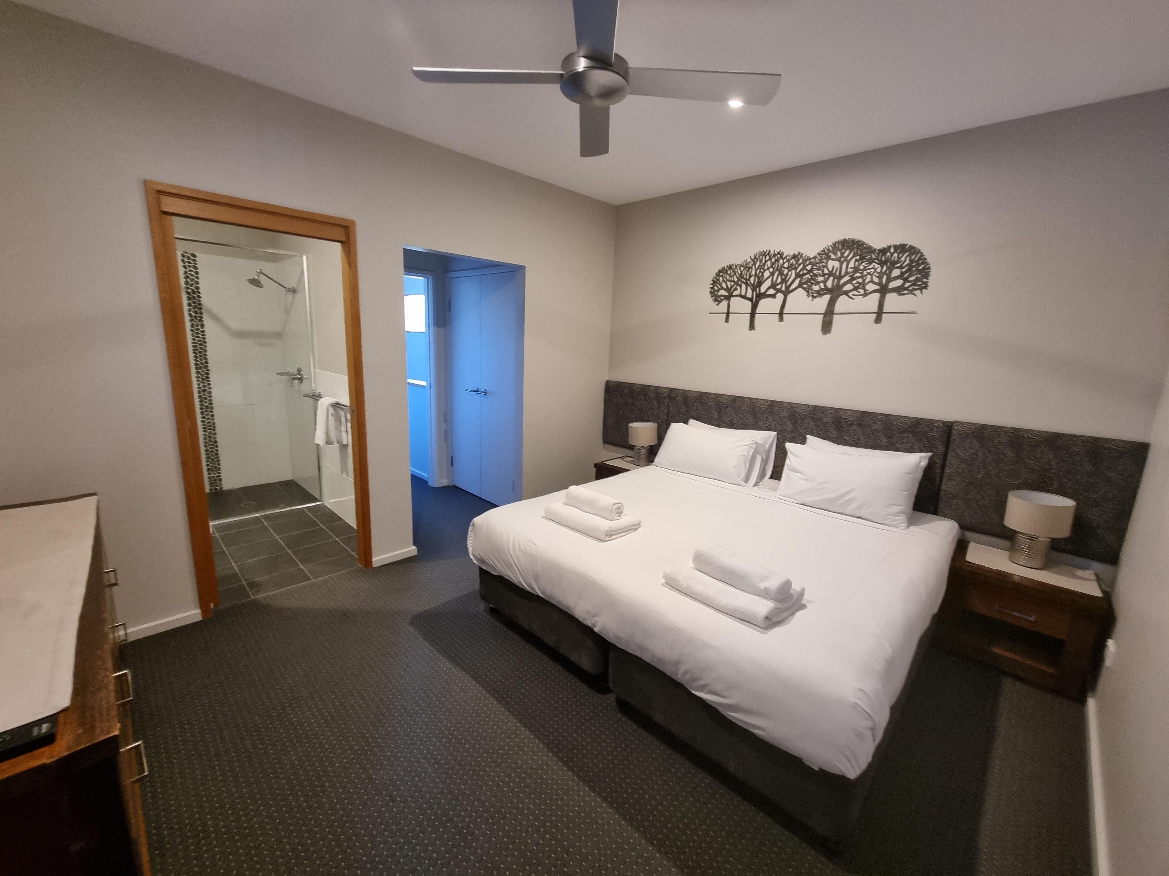 A hotel room with a bed, a nightstand, a ceiling fan, a shower area, and a wall art piece.
