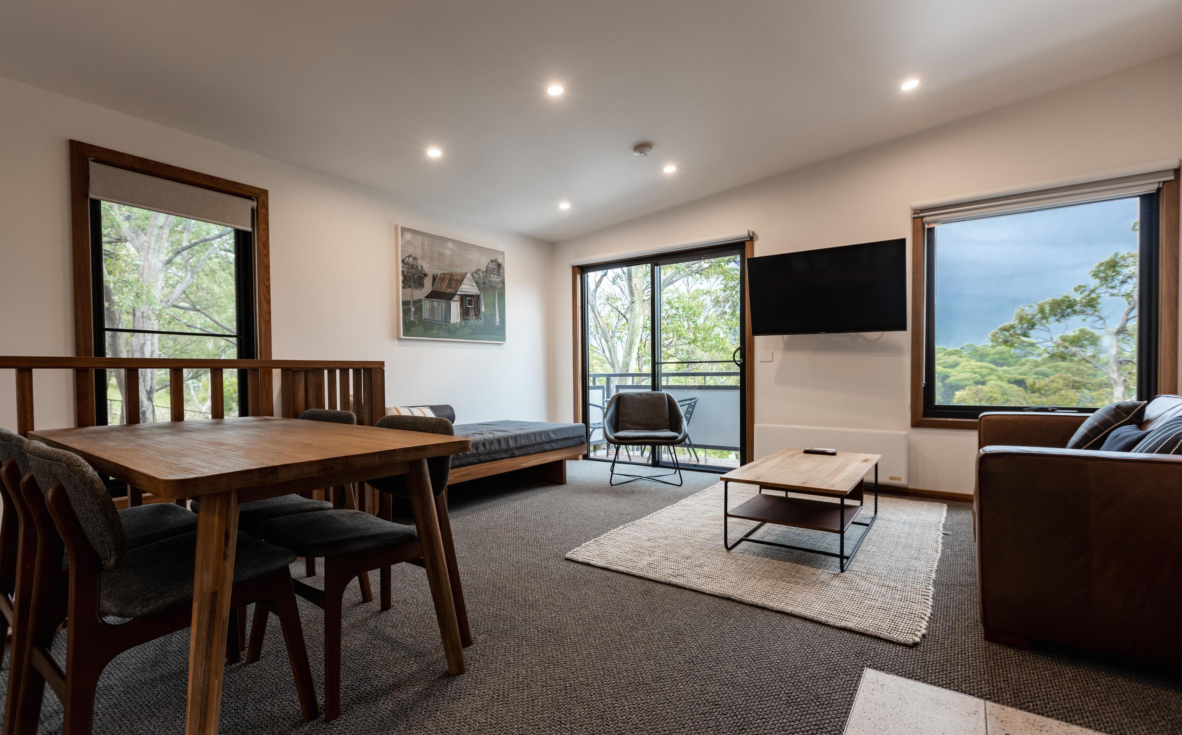 A modern living room with a wooden dining table, chairs, a sofa, a TV mounted on the wall, and large windows offering a view of the outdoors.