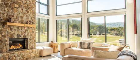 A spacious living room with a stone fireplace, large windows, and a view of a green landscape.