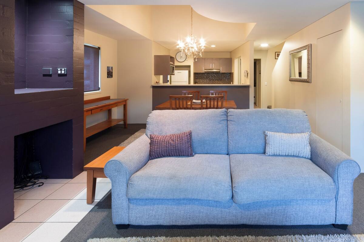A modern living room with a grey sofa, wooden dining table, and a kitchen in the background.