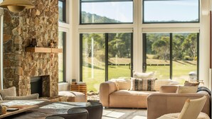 A spacious living room with a stone fireplace, large windows offering a view of a golf course, and comfortable seating. - Oaks Lake Crackenback Resort (Crackenback)