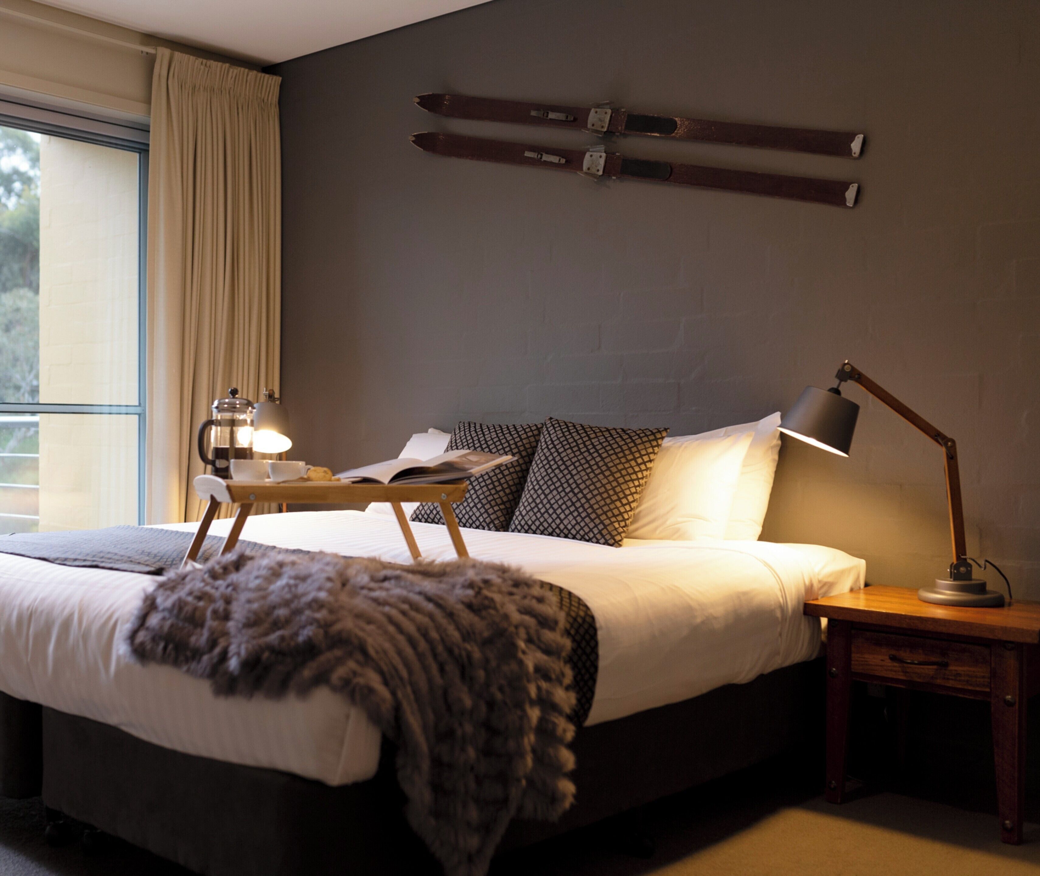 A modern bedroom with a large bed, a bedside table with a lamp, a window with curtains, and a wall-mounted ski rack.