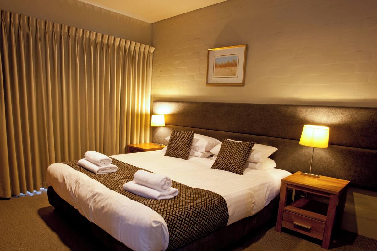 A hotel room with a large bed, two bedside tables, a headboard, a framed picture on the wall, and a window with curtains.