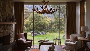 A cozy room with a stone fireplace, leather armchairs, a wooden coffee table, and a large window offering a view of a forested landscape. - Oaks Lake Crackenback Resort (Crackenback)
