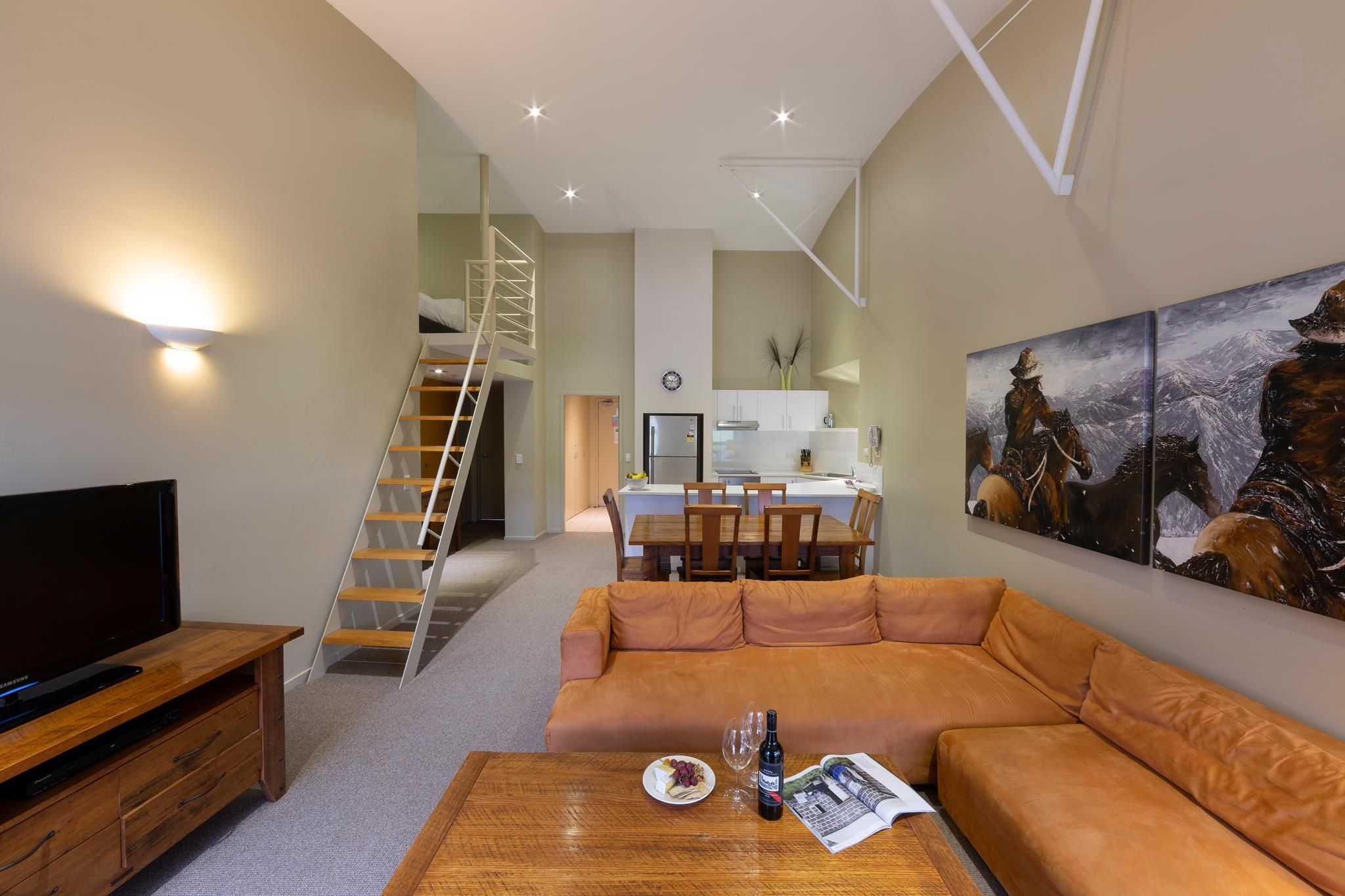 A modern living room with a sofa, coffee table, and a flat-screen TV. A staircase leads to a kitchen area with a dining table and chairs.