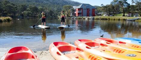Kayaks lined up on a sandy shore by a calm lake with a house in the background.