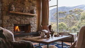 A cozy living room with a stone fireplace, a comfortable chair, a coffee table, and a view of a forested landscape through large windows. - Oaks Lake Crackenback Resort (Crackenback)