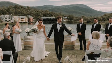 A wedding ceremony by a lake with guests seated on white chairs, a bride and groom walking down the aisle, and a backdrop of houses and hills.
