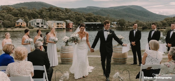 A wedding ceremony by a lake with guests seated on white chairs, a bride and groom walking down the aisle, and a backdrop of houses and hills. - Oaks Lake Crackenback Resort (Crackenback)