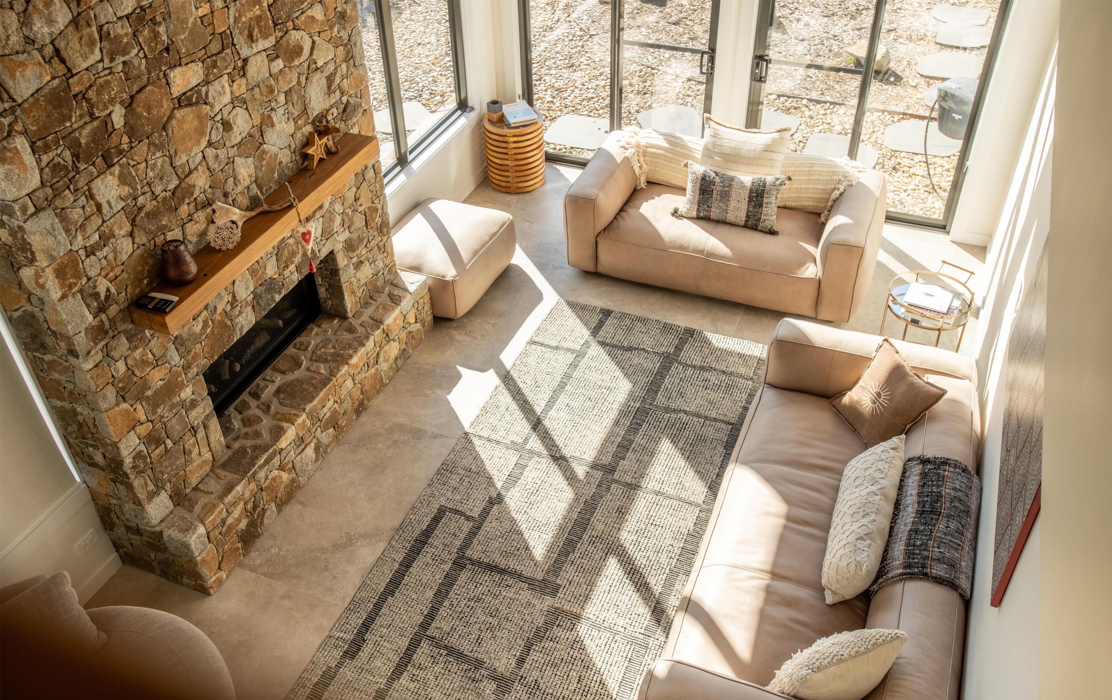 A cozy living room with a stone fireplace, a beige sofa set, and a large window offering a view of the outdoors.