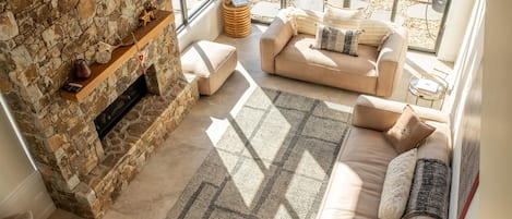 A cozy living room with a stone fireplace, a beige sofa set, and a large window offering a view of the outdoors.