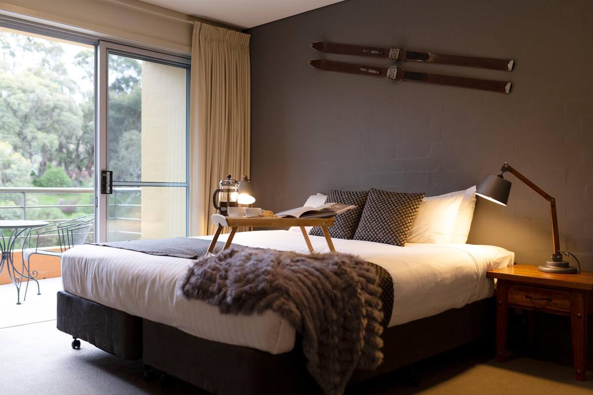A modern bedroom with a large bed, a bedside table with a lamp, a small table with a lamp, and a view of the outdoors through a sliding door.