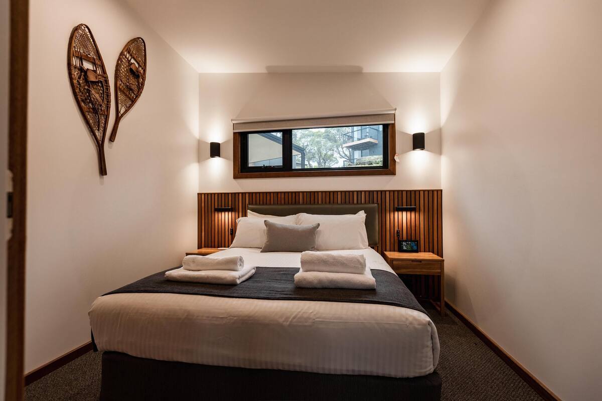 A neatly made bed with white linens and two towels, a wooden headboard, a window with blinds, and wall-mounted lights.