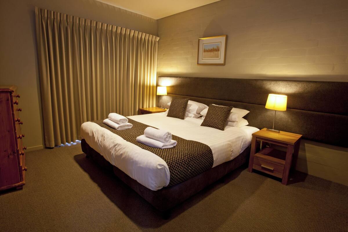 A hotel room with a large bed, two bedside tables with lamps, a wooden nightstand, and a large window with curtains.
