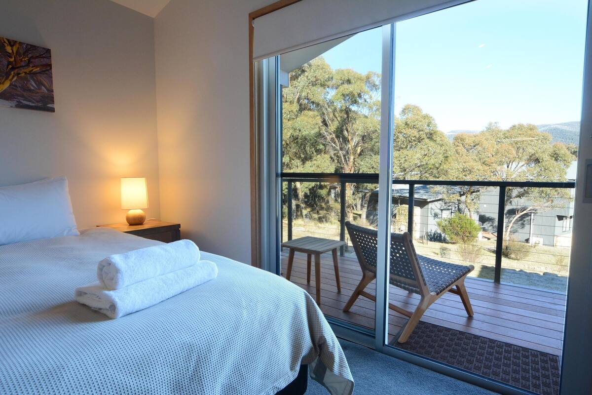 A bedroom with a large bed, a nightstand, a lamp, and a balcony with a chair and table.