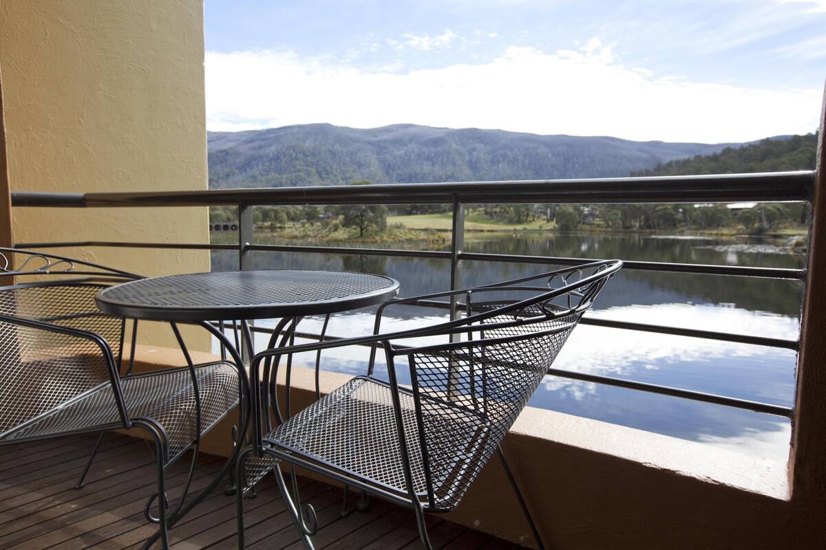 A balcony with a metal table and chairs, overlooking a lake and mountains.