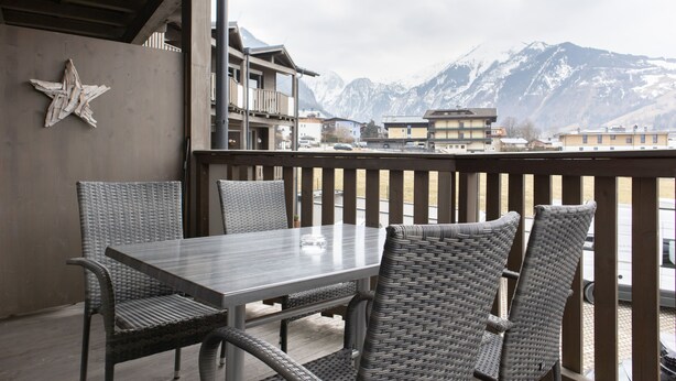 En balkon med fletstole og et bord, med udsigt til et bjergrigt landskab med bygninger.