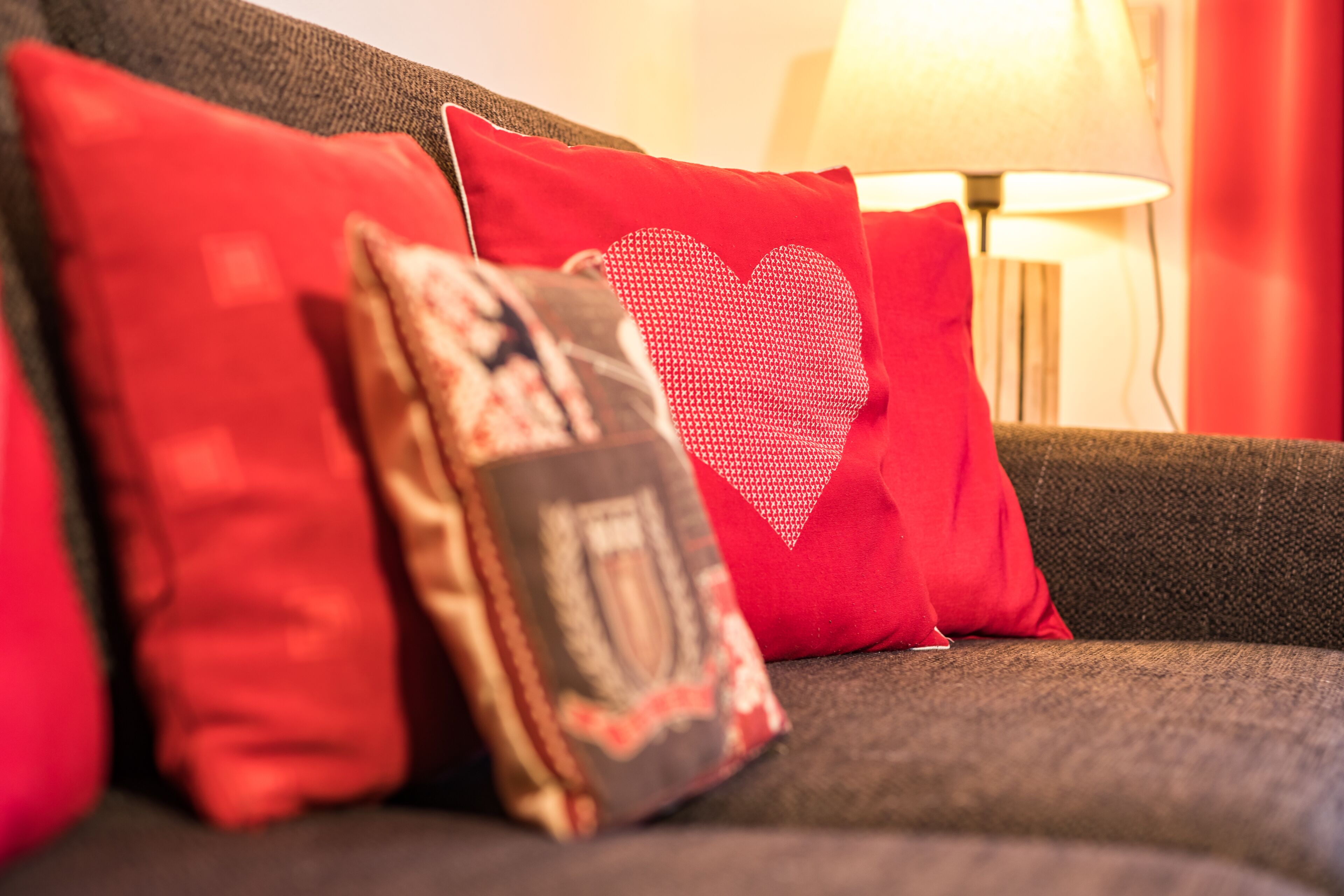 A sofa with red and patterned cushions, a heart-shaped cushion, and a lamp in the background.