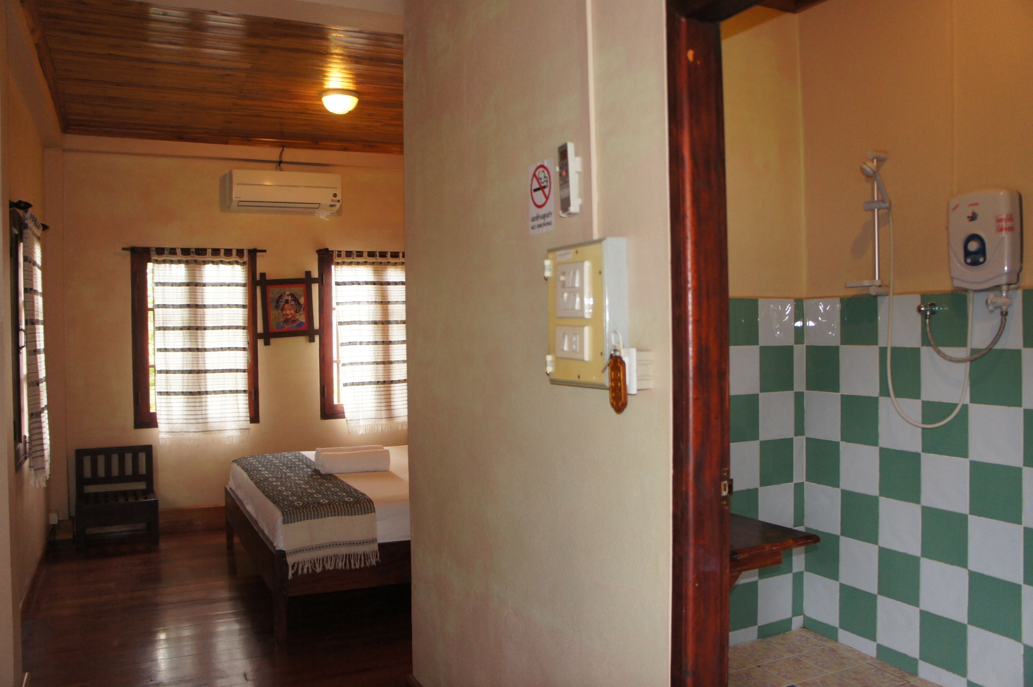 Una habitació amb llit, una panxa, una finestra amb cortines, un aparell d'aire condicionat, una làmpada penjada a la paret i un bany amb dutxa i dispensador de sabó.