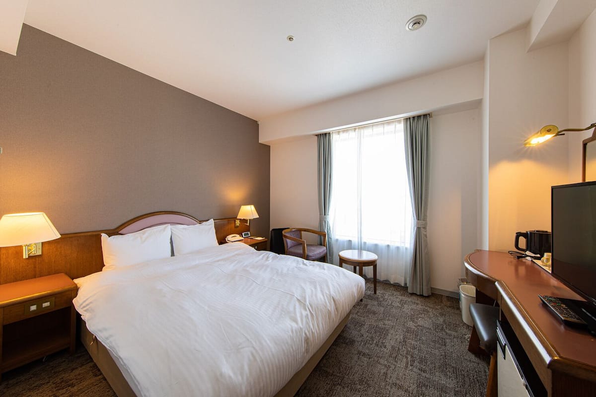 Ein Hotelzimmer mit Bett, Nachttischlampen, einem Schreibtisch mit Fernseher, einem Sessel und einem kleinen Tisch.