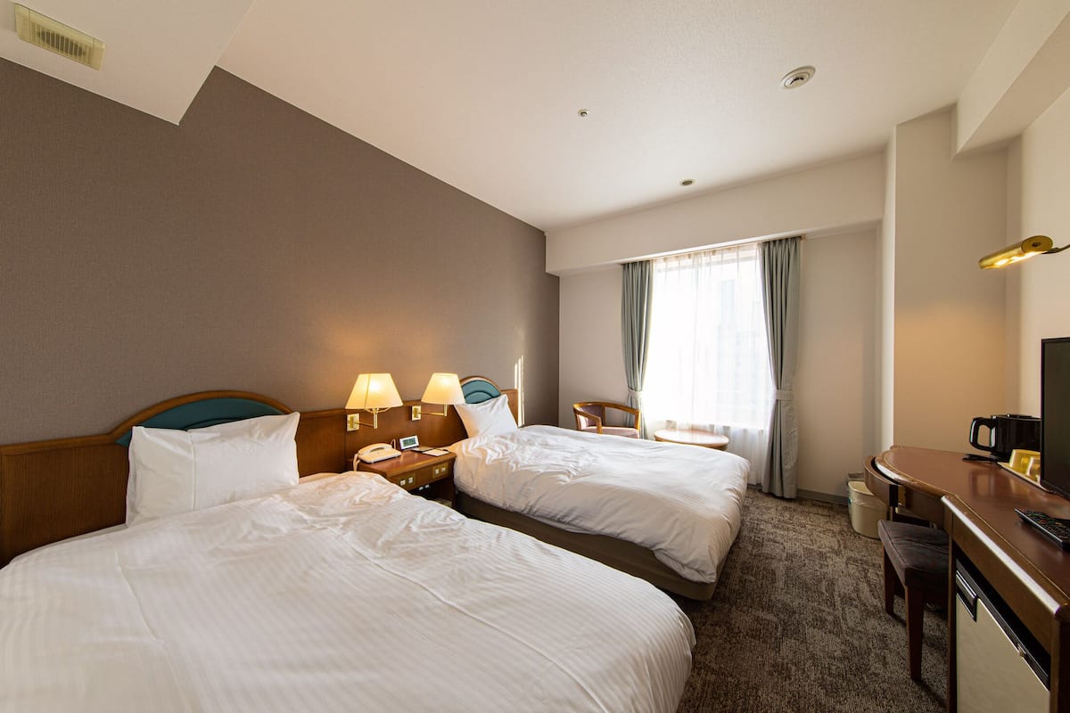 Ein Hotelzimmer mit zwei Betten, einem Schreibtisch, einem Stuhl, einem Fernseher und einem Fenster mit Vorhängen.