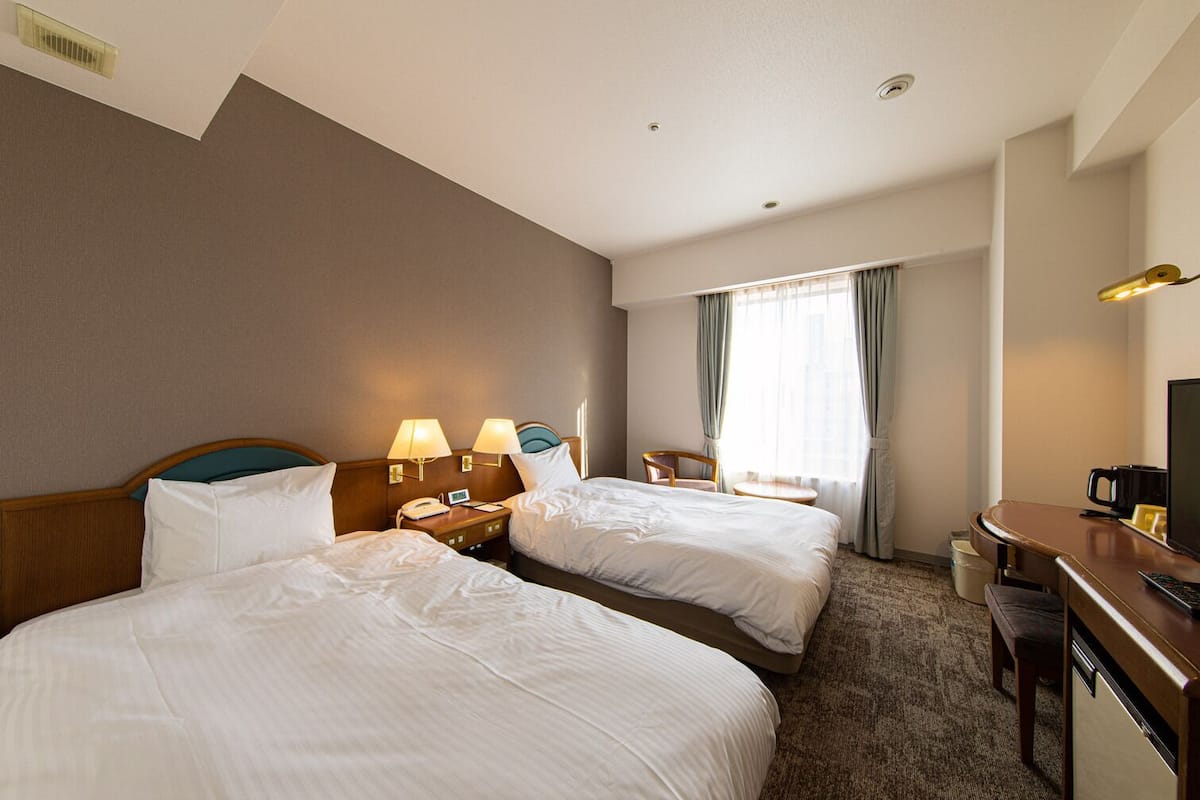 Ein Hotelzimmer mit zwei Betten, einem Schreibtisch, einem Stuhl, einer Schreibtischlampe, einem Fernseher und einem fenster mit Vorhängen.