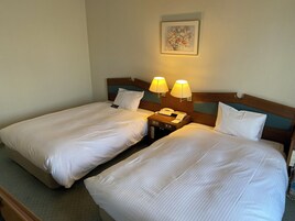 Quarto de hotel com duas camas, cada uma com roupa de cama branca, cabeceira de madeira e duas luminárias de cabeceira. Há uma obra de arte emoldurada na parede acima das camas.