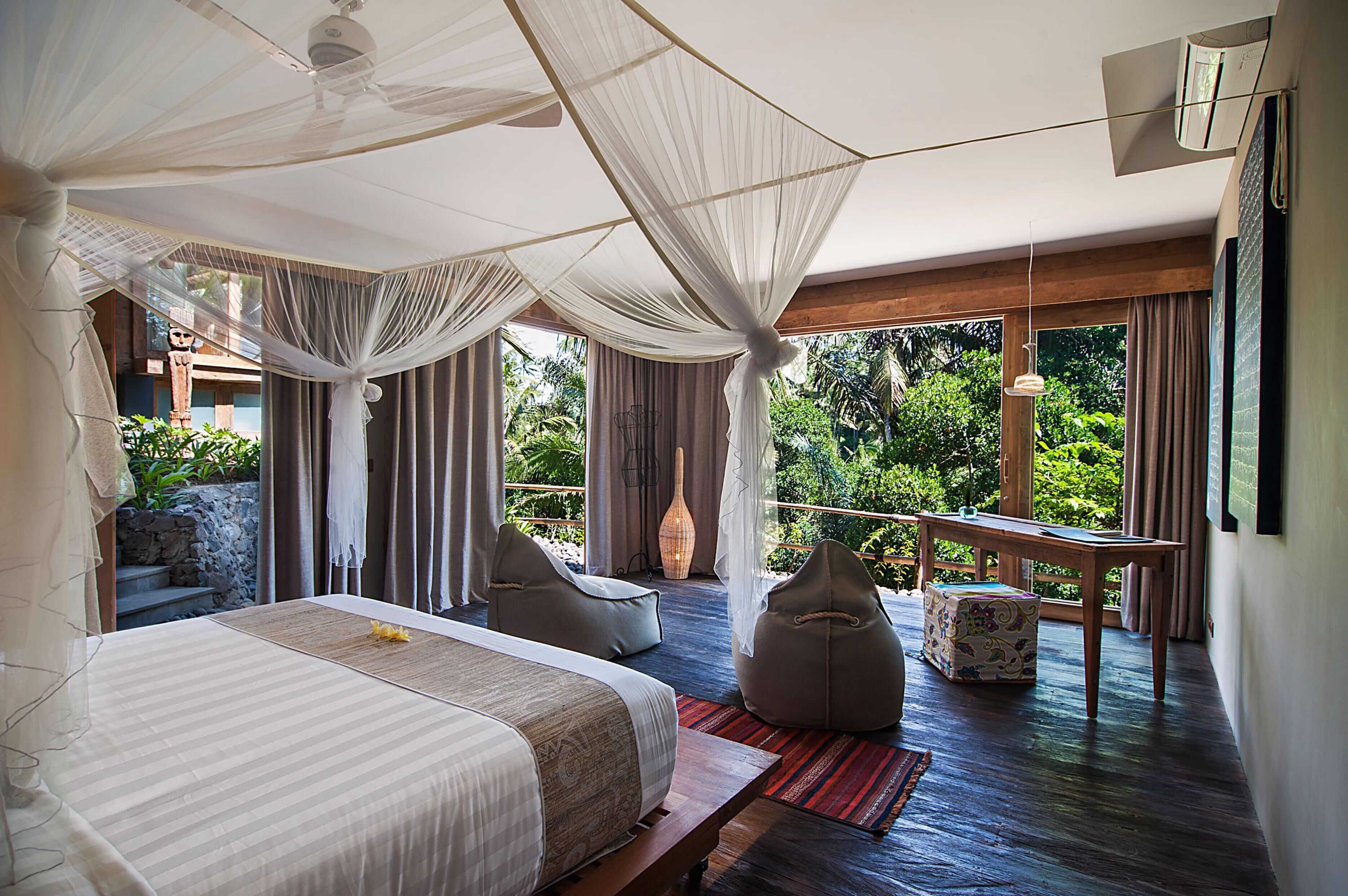 A bedroom with a large bed, a sitting area with chairs, a small table, and a view of lush greenery.