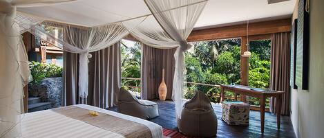 A bedroom with a large bed, a sitting area with chairs, a small table, and a view of lush greenery.