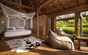 A bedroom with a bed, a canopy, a cowhide rug, a chair, and a view of greenery through large windows. - Blue Karma Dijiwa Ubud (Ubud)