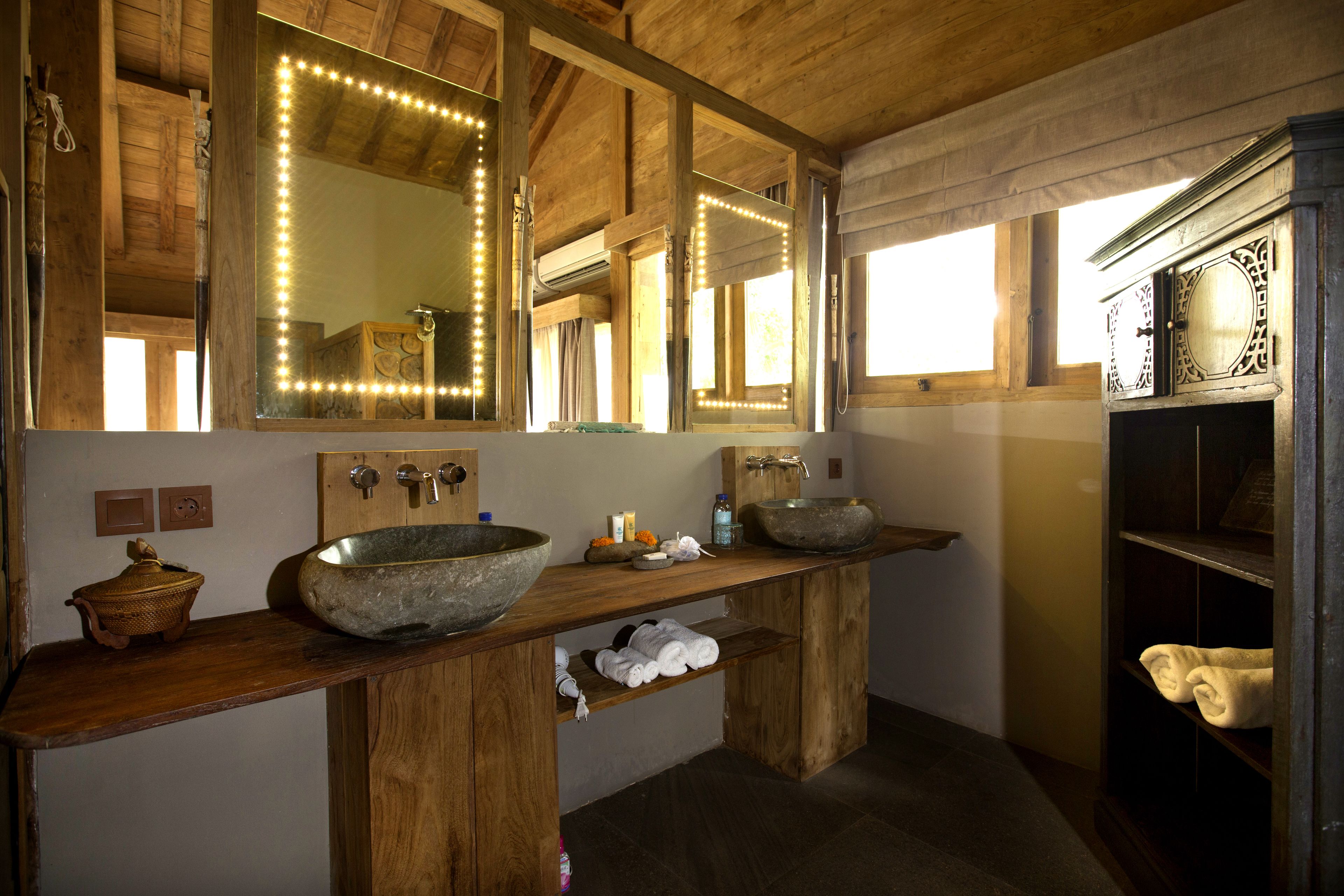 A rustic bathroom with wooden beams, a large mirror with lights, two stone sinks, and a wooden cabinet with towels.