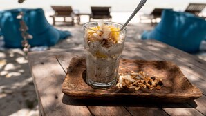 Un vaso de cereal con una cuchara sobre una tabla de madera, colocado en una playa con sillas y vista al mar.