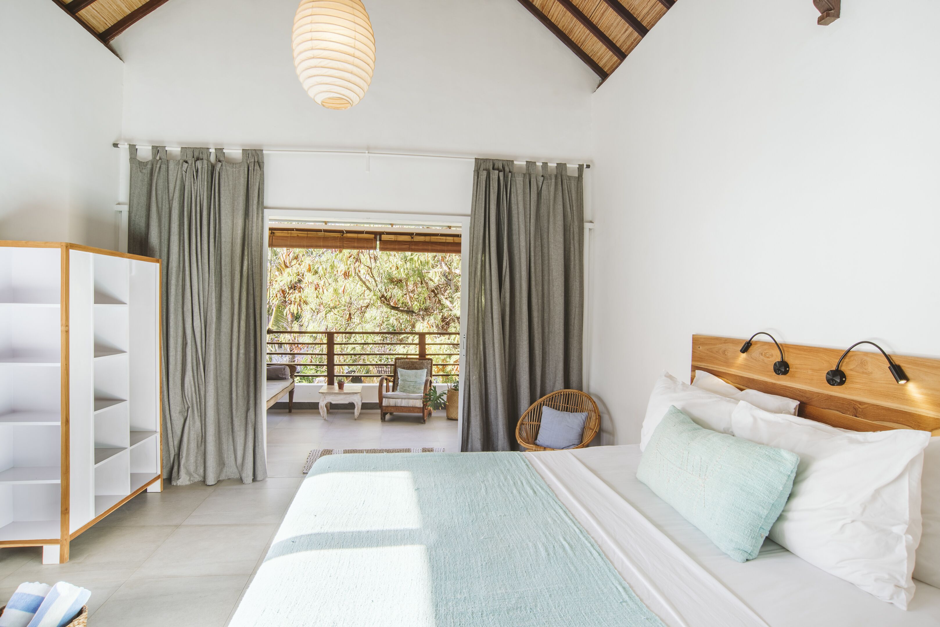 A bedroom with a bed, a wardrobe, a balcony with a view of trees, and a hanging light fixture.