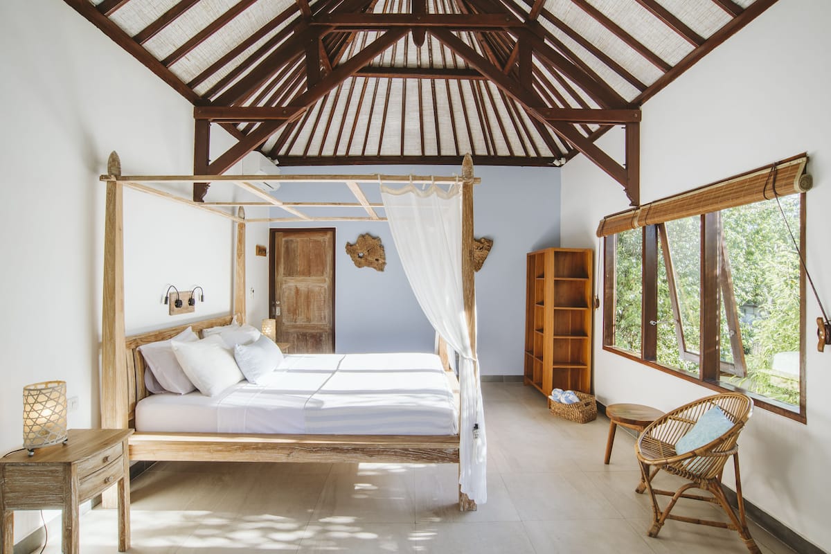 A bedroom with a wooden four-poster bed, a wooden wardrobe, a wicker chair, and a large window with a view of greenery.