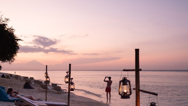 Una playa con sillones de madera y faroles, una persona tomando una foto, y una puesta de sol al fondo.