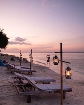 A beach with wooden lounge chairs and lanterns, a person taking a photo, and a sunset in the background. - Karma Kayak (Gili Trawangan)