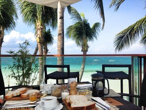 Una mesa de comedor dispuesta con pan, café y té, con vista a una playa con palmeras y el mar.