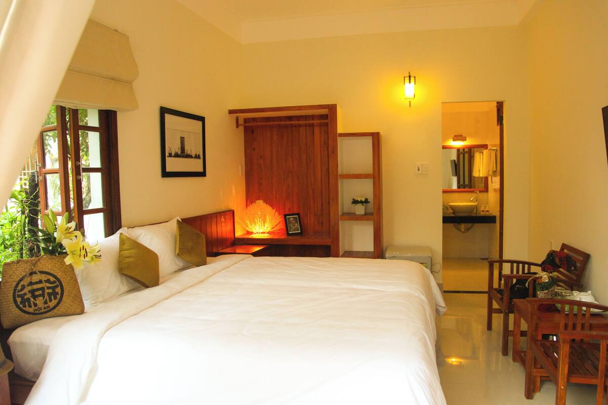 A hotel room with a bed, a nightstand with a lamp, a wooden headboard, a dining table with chairs, and a bathroom visible through an open door.